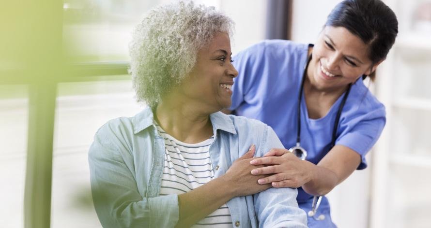 Senior woman in wheelchair receives medical help for injury A senior woman sits in a doctor's office in a wheelchair. She smiles back at a cheerful mid adult female doctor behind her. The doctor puts her hand on hers.